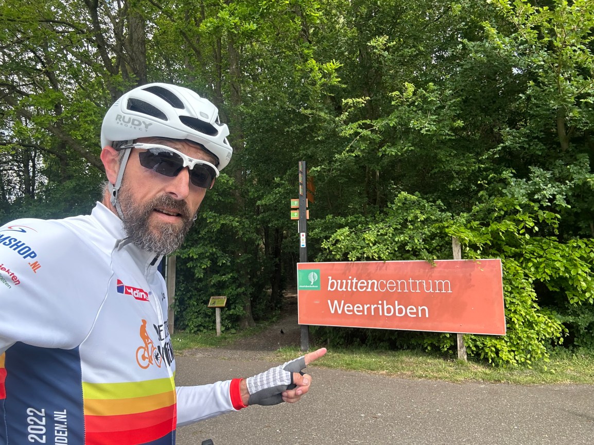 Robert Baarda in fietskleding van De Fietsende Vrienden wijzend naar een bord van Buitencentrum Weerribben