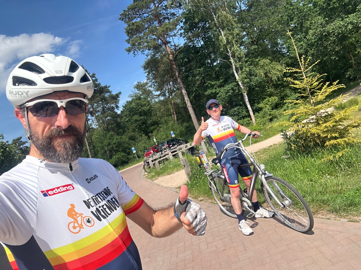 Robert Baarda op de voorgrond en Age Yska op zijn fiets in de achtergrond poseren in hun De Fietsende Vrienden fietskleding met hun duim omhoog bij het bezoekerscentrum van 't Leesten in Ugchelen